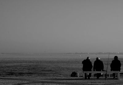 Three people sitting on bench next to sea against clear sky