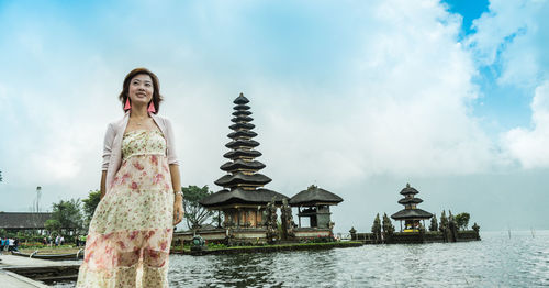 Low angle view of woman looking away while standing by lake against temple and sky
