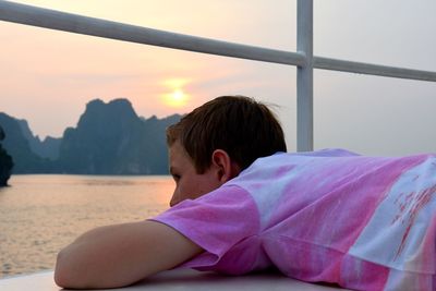 Boy leaning at observation point by sea against sky during sunset