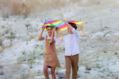 A girl and a boy are carrying a bright kite with their hands raised high above their heads.