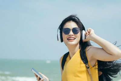 Portrait of young woman using mobile phone while listening to music against sea