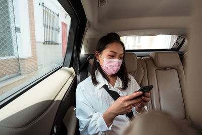 Ethnic female passenger with fastened seat belt using the cell phone while riding in protective mask on backseat in taxi