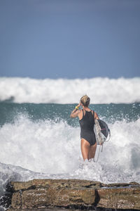 Rear view of woman with surfboard standing on rock at sea