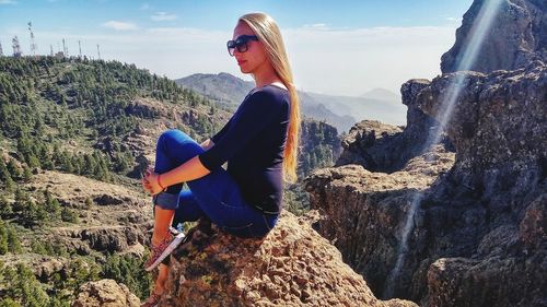 Side view of young woman sitting on rock against sky
