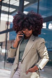 Positive black businesswoman in suit with curly hair holding hand in pocket and answering phone call while standing on modern city street before work