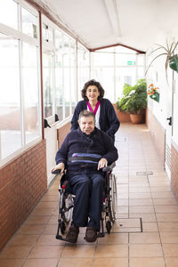 Smiling female caregiver assisting disabled man sitting on wheelchair in corridor