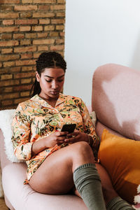 Modern african american female in casual wear sitting on comfortable sofa and browsing smartphone while resting in cozy living room with loft interior