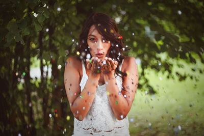 Portrait of young woman blowing confetti while standing against trees in park