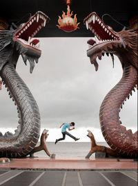 Man jumping at beach seen through dragon statues against sky