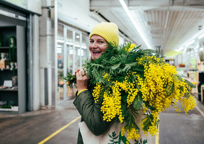 Positive woman in yellow cap and large bouquet of mimosa flowers at flower market