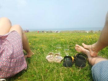 Low section of couple resting by champagne flutes on grassy field against clear sky