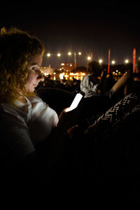 Side view of woman sitting at illuminated park against sky at night