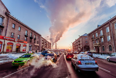 Cars on city street amidst buildings against sky during sunset