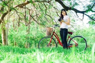 Portrait of young woman photographing while standing with bicycle on grassy field in forest