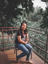 Portrait of young woman sitting on railing against trees