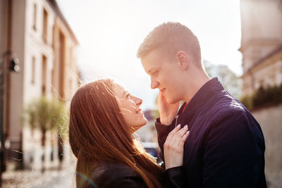 Couple looking at each other while standing amidst buildings against sky