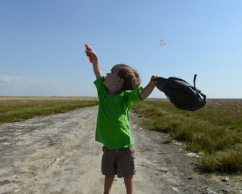 Boy flying kite while standing on road amidst grassy field against sky during sunny day