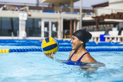 Smiling young woman with ball swimming in pool on sunny day