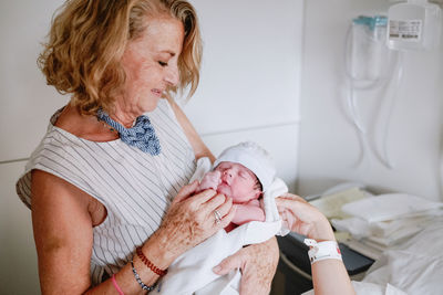 Happy mature female greeting newborn grandchild in hospital ward while young mother lying in bed and receiving medical treatment