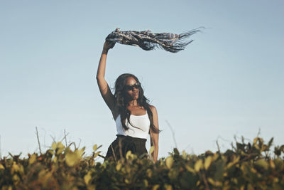 Low angle view of woman with arm raised holding scarf against sky