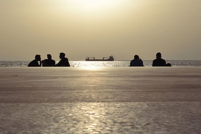 Silhouette men sitting at beach against sky during sunset