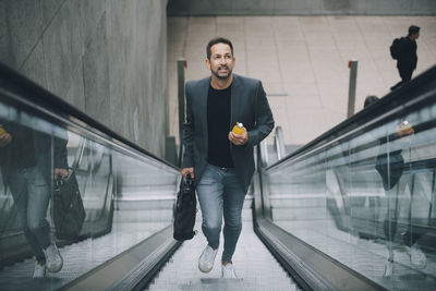 Confident businessman holding drink while moving on escalator in subway