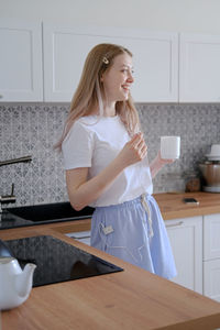 A cute girl of 15-18 years old stands in the kitchen with a mug in her hands