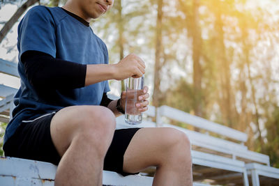 Midsection of athlete holding water bottle while sitting on bench at park