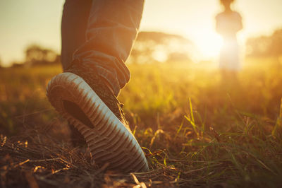 Low section of man walking on grassy field during sunset