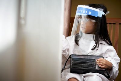 Portrait of sad little girl with face protective mask looking through the window at home quarantine