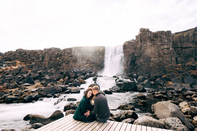 Man sitting on rock by waterfall against sky