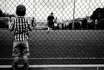 Rear view of boy standing by chainlink fence against sky
