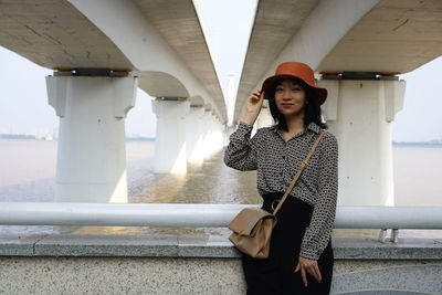 Portrait of beautiful young woman standing against bridge