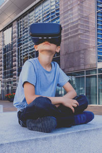 Boy wearing virtual reality simulator while sitting against building in city