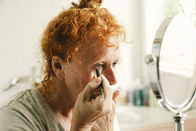 Senior woman looking in mirror and applying eyeliner at home