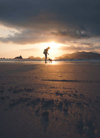 Silhouette woman standing on beach with dog against sky during sunset