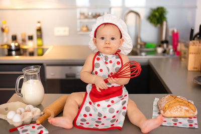 Full length of cute girl holding wire whisk sitting on table at kitchen