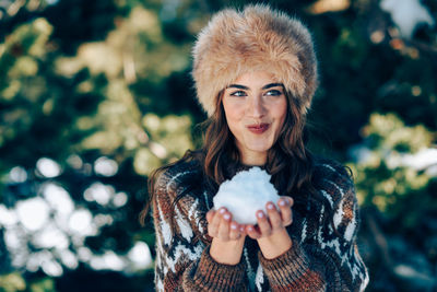 Beautiful woman holding snowball while standing against tree