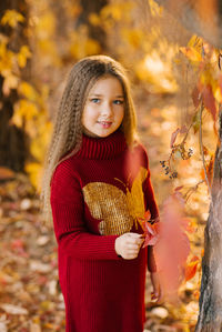 Children girl in a burgundy knitted tunic stands near a tree in a park with yellow and red leaves