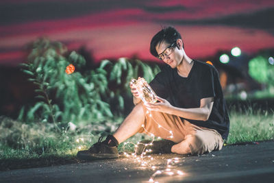 Young man holding jar and illuminated string lights while sitting on road at night