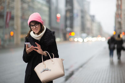 Woman using smart phone while standing in city during winter