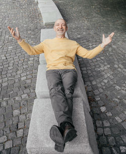 Senior man in yellow sweater reclining on an urban bench with arms open wide