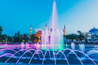 Illuminated fountain against buildings in city at dusk