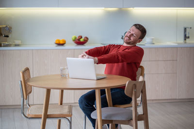 Positive freelancer man smiling with closed eyes stretching sitting on kitchen with laptop at home