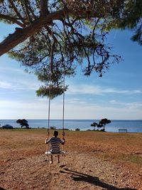 Rear view of a boy sitting on a swing on beach against sky