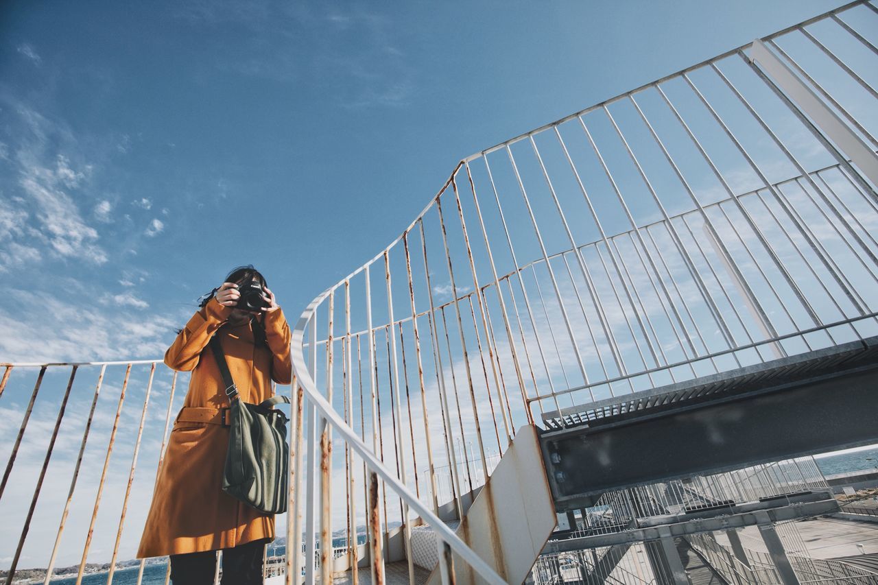 real people, sky, one person, architecture, railing, lifestyles, low angle view, leisure activity, standing, built structure, clothing, nature, cloud - sky, young adult, day, connection, adult, young women, front view, bridge - man made structure, outdoors, warm clothing