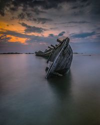 Damaged boat in sea against sky during sunset