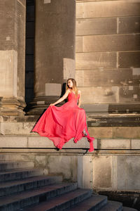 Full length of young woman in pink evening gown sitting against columns