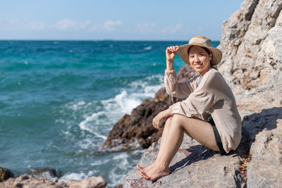 Full length of smiling woman sitting on rock at beach against sky