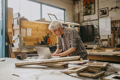 Focused senior male carpenter working on laptop at repair shop